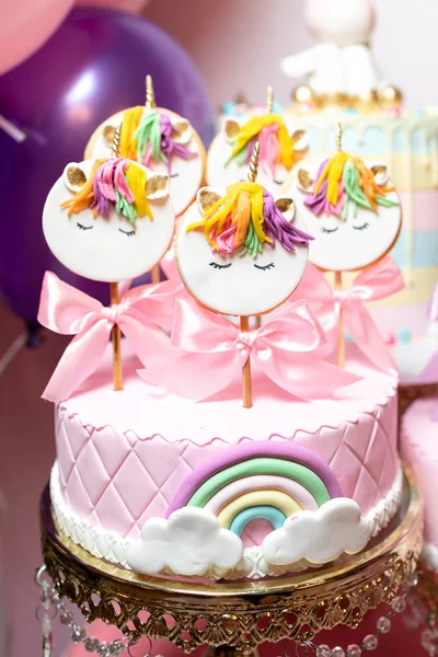 Pink cake with marzipan, with a rainbow and white clouds, and cookies on sticks with the image of a unicorn.