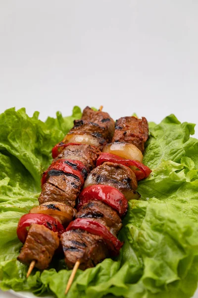 Bitar av stekt kött och grönsaker på spett som ligger på en tallrik på ett sallads blad på vit botten. — Stockfoto