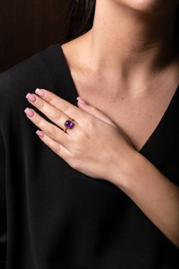 Ortasında mor taş olan altın kadın yüzüğü parmağında kare şeklinde, siyah bir arka planda..