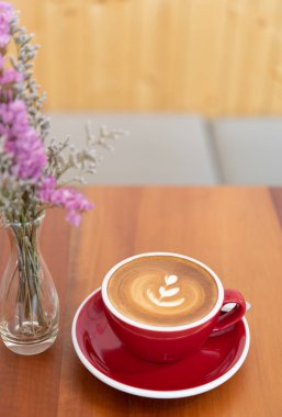 Ahşap masa ve çiçek vazo üzerinde latte sanat ile Kırmızı sıcak kahve fincanı