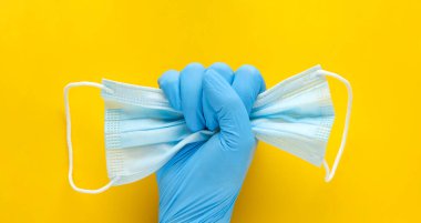Sarı arka planda tek kullanımlık tıbbi maske tutan eldivenli bir el. Kişisel koruyucu ekipman eksikliğine öfke, PPE, sağlık çalışanları konseptine.