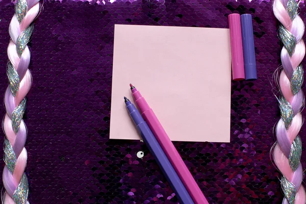 Pink sheet of note paper and pink and purple felt-tip pens on a dark pink background fabric with sequins shines and shimmers and a pigtail of shiny threads, free space for records