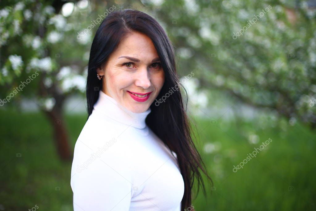 Young smiling woman with long black hair in a white turtleneck spring flowering lilac and apple trees