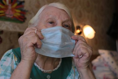 Mavi tıbbi maske, yaşlı, gri saçlı bir büyükannenin elinde. Virüslere karşı korumak için solunum cihazı takın.