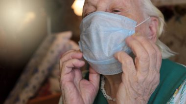 Mavi tıbbi maske, yaşlı, gri saçlı bir büyükannenin elinde. Virüslere karşı korumak için solunum cihazı takın.