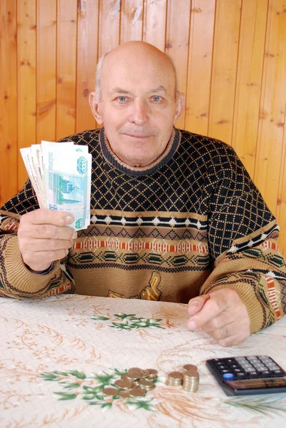 The grandfather at the dacha sits at a table with the calculator and holds paper notes in hand. The elderly person sits at a table with the calculator and shows the money holding in hand.