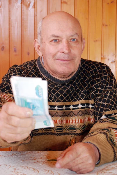 The grandfather at the dacha sits at a table and stretches paper notes. The elderly person sits at a table and stretches the money holding in hand.