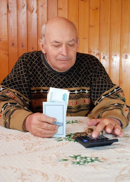 The grandfather at the dacha sits at a table with the calculator, money and the savings book and considers pension. The elderly person sits at a table with the calculator, paper notes and coins and considers pension.