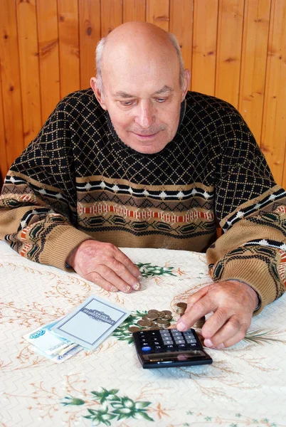 The grandfather at the dacha sits at a table with the calculator, money and the savings book and considers pension. The elderly person sits at a table with the calculator, paper notes and coins and considers pension.