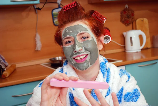 The young girl with red hair in hair curlers with a clay face pack in kitchen does manicure