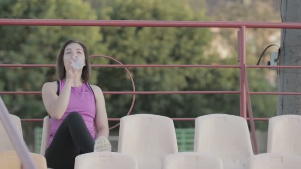 Красивая фитнес-девушка пьет воду из бутылки после тренировки на стадионе — стоковое видео