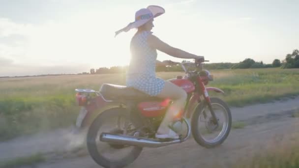 Uma jovem feliz em um chapéu monta uma motocicleta retro e desfruta de um passeio em uma estrada de terra entre os campos e prados — Vídeo de Stock