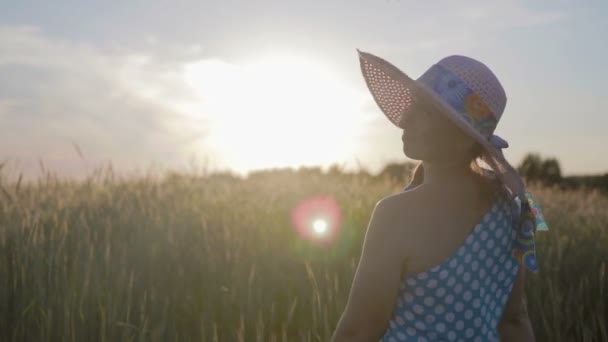 Retrato de uma mulher feliz em um vestido leve e chapéu. A menina caminha no campo com plantas de cereais ao pôr do sol — Vídeo de Stock