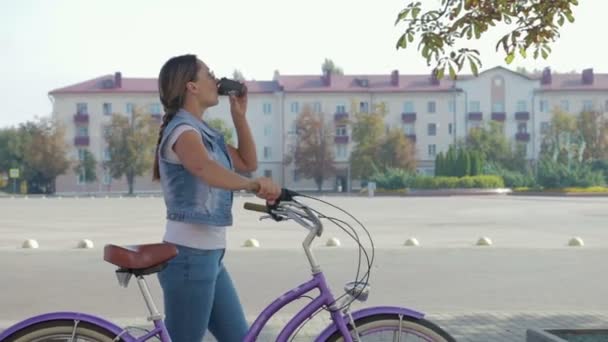 Retrato de una chica joven y feliz que pasea por la ciudad con una bicicleta y bebe café — Vídeos de Stock