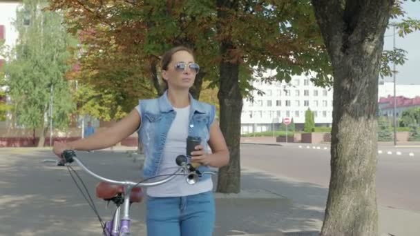 Porträtt av en ung, glad tjej som går runt i staden med cykel och dricker kaffe — Stockvideo