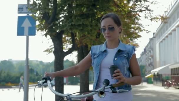 Porträtt av en ung, glad tjej som går runt i staden med cykel och dricker kaffe — Stockvideo