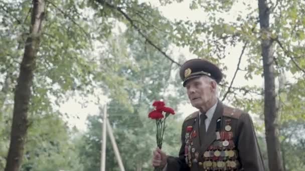 Podeszłym wieku siwy weteranów Wielkiej Wojny Ojczyźnianej i Ii wojny światowej w mundurze z wielu odznaki i zamówień trafia do pomnika. Dziadek jest w rękach czerwone goździki — Wideo stockowe