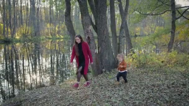 Madre e hijo en ropa elegante caminar en el bosque de otoño, cerca del lago, el reflejo de los árboles se puede ver en el lago — Vídeos de Stock