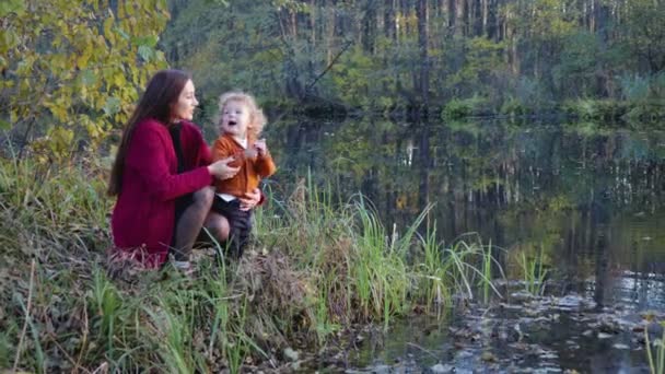 Mor och barn sitter, prata och kramas nära sjön mot höstlandskap — Stockvideo