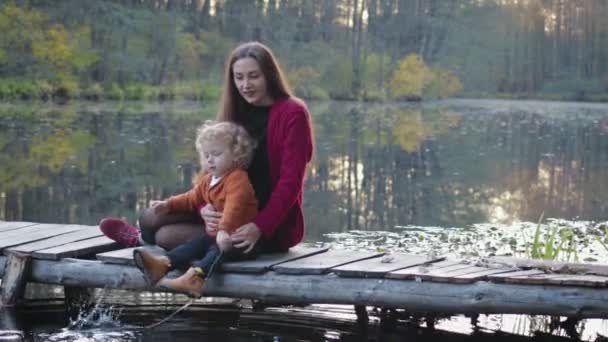 Madre e figlio si siedono su un ponte di legno sul lago e godono della splendida vista. Il concetto di famiglia — Video Stock