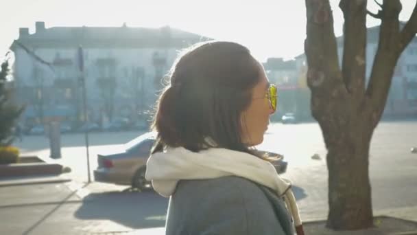 Porträtt av en ung flicka på sida, hon går runt i staden i en kappa och glasögon — Stockvideo