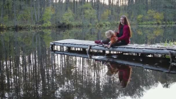 Schöne und stilvolle Mutter und Sohn sitzen auf der Brücke am See im Park und bewundern die herbstliche Landschaft. — Stockvideo