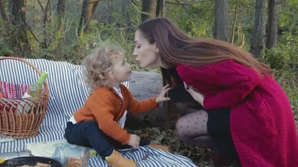 Счастливая мать целует своего ребенка на пикнике в лесу и дает ему телефон — стоковое видео