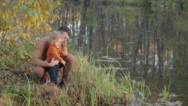 Famiglia giovane ed elegante seduta sull'erba sulla riva del fiume, papà e figlio giocano e si divertono — Video Stock
