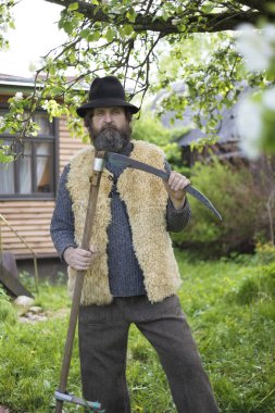 Yazın köyde sakallı ve şapkalı yetişkin bir adam elinde tırpanla evinde poz veriyor.