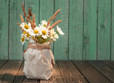 Beyaz papatya çiçekleri, kraft kağıdından bir vazoda kurumuş kulaklar, köhne ahşap bir arka planda.