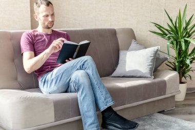 Genç adam parlak bir odada dinleniyor. Kanepede oturuyor ve kitap okuyor.