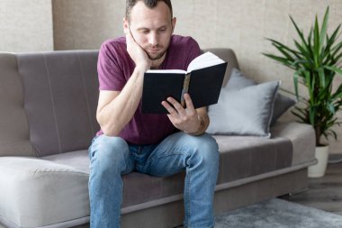 Genç ve sıkılmış bir adam kanepede otururken kitap okuyor.