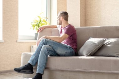 Genç adam, parlak bir odada, kanepede oturuyor ve pencereden dışarı bakıyor.