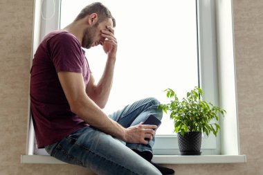 Tişörtlü ve kot pantolonlu genç adam büyük bir pencerenin pervazında oturuyor. Üzülerek başını eğiyor.