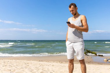 Genç adam sahilde denize karşı duruyor ve akıllı telefona bakıyor.