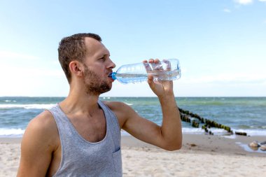 Genç adam deniz kıyısındaki bir şişeden su içiyor.