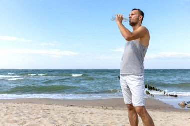 Genç adam deniz kıyısındaki bir şişeden su içiyor.