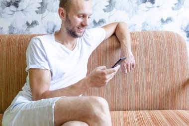 Genç adam bir kanepede otururken akıllı telefonun ekranına bakar.