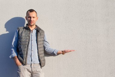 Gri kot pantolon, mavi kot gömlek ve yelek giyen yakışıklı genç adam parlak duvarın yanında duruyor.