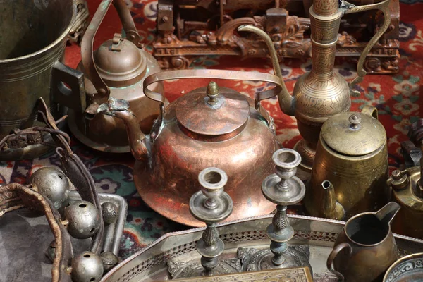 On an old carpet there are different old metal things. In the centre there is a copper teapot.
