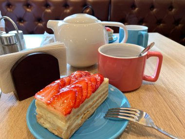 Mavi tabakta çilekli kek. Açık kahverengi masa beyaz su ısıtıcısı, pembe ve mavi kupa.