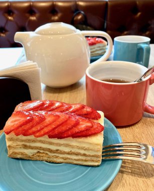 Mavi tabakta çilekli kek. Açık kahverengi masa beyaz su ısıtıcısı, pembe ve mavi kupa.