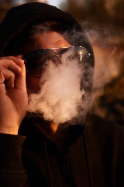 Güneş gözlüklükteki adam elektronik sigara içiyor. Vaper hipster duman buharlaştırıcı ve duman bulutu exhals