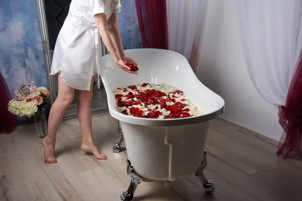 Chica en una túnica de satén blanco espolvorea los pétalos de rosas escarlata en el baño blanco vintage —  Fotos de Stock