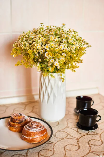 明るい色のキッチンで朝、小さなヒナギクの花束、黒いカップとシナモンロールのハーブティー — ストック写真