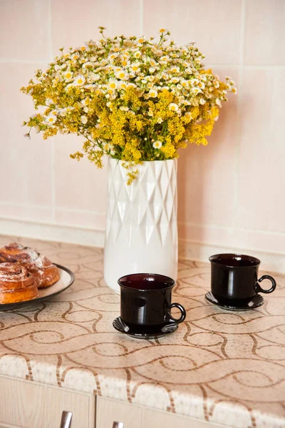明るい色のキッチンで朝、小さなヒナギクの花束、黒いカップとシナモンロールのハーブティー — ストック写真