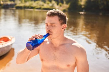 çıplak gövdeile kaslı bir adam sıcak bir günde bir nehir veya gölün kıyısında duruyor ve soğuk su içiyor