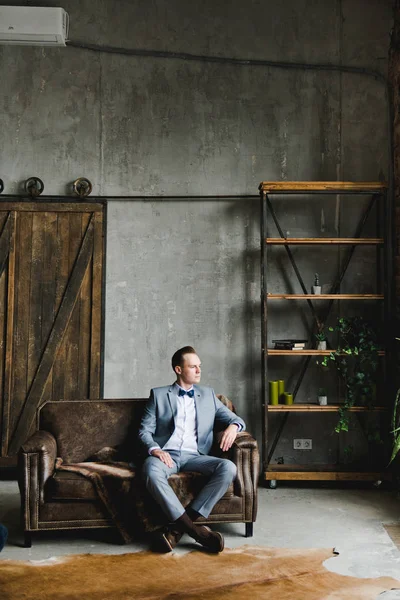 Retrato de un hombre. El novio en un traje gris, camisa blanca y corbata de lazo está sentado en un sofá de cuero marrón. Habitación en estilo loft . — Foto de Stock