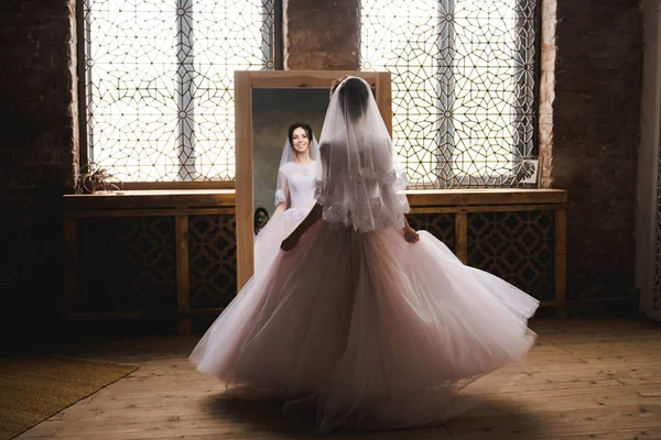 La novia se reúne por la mañana cerca del espejo. La novia se reunirá por la mañana. Elegante vestido de novia rosa. La novia está girando en un vestido cerca del espejo . — Foto de Stock