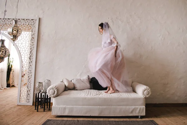 La novia salta al sofá. La novia se reunirá por la mañana. Elegante vestido de novia rosa . — Foto de Stock